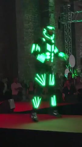 A person in a black costume with illuminated red and yellow lights dances on a red stage in front of a stone wall and an audience. The costume has geometric patterns of lights on the chest, arms, and legs. The dancer moves dynamically, with arms raised and legs bent. The environment appears to be indoors, possibly an event space or performance venue, with stage lighting and a truss structure visible in the background. The audience is seated and observing the performance.