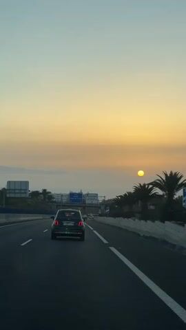 The video shows a car driving on a highway during sunset. The sun is a bright yellow orb near the horizon, casting a warm glow across the sky. The sky transitions from a pale blue at the top to a soft orange and yellow as it approaches the sun. Palm trees line the right side of the highway, silhouetted against the colorful sky. Road signs are visible overhead, indicating directions to different locations. Another car is driving in front of the camera vehicle. The road is paved and has white lane markings. The overall scene depicts a serene and picturesque drive as the day comes to a close.