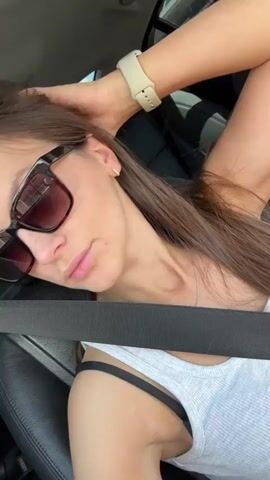 A young woman with long brown hair and sunglasses is sitting in the passenger seat of a car. She is wearing a white tank top and a black bra is visible. She is holding her left arm up, resting her hand on her head. The seatbelt is across her chest. The interior of the car is visible, including the black leather seats and a window with a view of buildings and trees. The woman is looking towards the camera.