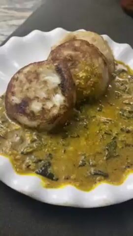 The video shows a plate of food. There are three round, slightly browned, pancake-like objects stacked together on a white plate. The pancakes rest on a yellowish-brown sauce with green leafy vegetables mixed in. The plate has a ruffled edge. The background is blurred.