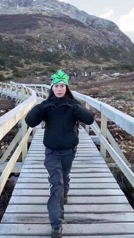 A person in hiking attire walks toward the camera on a wooden boardwalk, then the camera swings to show the boardwalk winding into a landscape of patchy vegetation, rocky ground, and mountains in the background. The person is wearing a black puffer jacket, gray hiking pants with white stripes, and a green beanie with a star. The boardwalk is gray and has white railings. Other people can be seen in the distance on the path. The mountains in the background are partially covered in snow and clouds. The sky is partly cloudy.