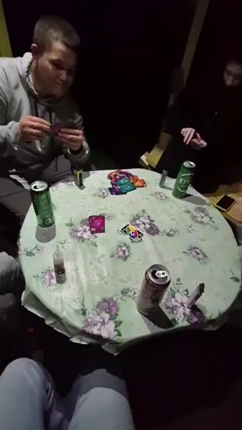 Four people are gathered around a round table covered with a floral tablecloth, playing a card game, possibly UNO, as seen from the visible cards. The setting appears to be outdoors or in a dimly lit indoor space. The table has several green cans, one opened soda can, and what appears to be a vape pen. The people are dressed casually. One person is holding a hand of cards, another is placing a card on the table, and the other two are also engaged in the game. The lighting is low, creating shadows.