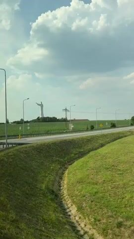 The video shows a landscape scene with a large statue of Jesus Christ in the background, situated on a hill overlooking a field of crops. The statue is white and appears to be made of stone or concrete, with its arms outstretched. In the foreground, there is a road with traffic signs, including a roundabout sign and a yield sign. The scene is set in a rural area, with green fields and trees. The sky is blue with scattered clouds, suggesting it is a bright and sunny day. The overall mood is peaceful and serene.

The video also captures some traffic-related infrastructure, such as guardrails and lampposts. The presence of a roundabout sign indicates that this is likely a junction or intersection. The statue of Jesus Christ is a prominent landmark, suggesting a possible tourist attraction or religious site. The video appears to have been filmed from a moving vehicle, likely a car or bus, given the slight motion blur and the perspective of the road.

While there are no people visible in the video, the scene evokes a sense of grandeur and spirituality due to the presence of the statue. The landscape suggests a location in a temperate climate, likely during the spring or summer months when the crops are growing. The vastness of the fields and the open sky contribute to a feeling of freedom and tranquility. The combination of natural beauty and religious symbolism creates a visually striking image.

The video does not contain any visible text, except for the road signs which are pictorial rather than text-based. The time of day is likely daytime, as indicated by the bright sunlight and the clear visibility of the landscape. The weather appears to be fair, with a mix of sun and clouds. The location cues, such as the style of the statue and the surrounding landscape, could potentially help identify the specific location, but without further information, it is difficult to pinpoint the exact geographical coordinates. However, the provided location context of Osogóra, Poland, offers a significant clue to the setting.
