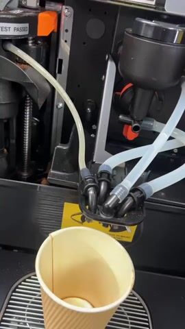 A close-up video shows coffee being poured from a machine into a brown paper cup. The machine has black and silver parts with several clear tubes attached to a black dispenser head. The coffee starts as a dark brown and lightens as it fills the cup, creating some bubbles. A yellow warning sticker is visible on the machine near the cup. The background consists of the machine's components.