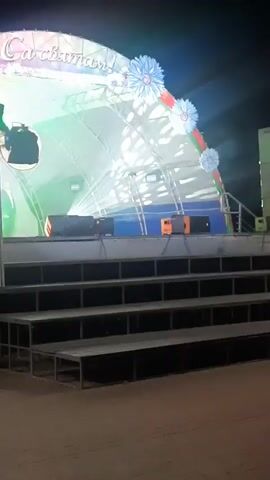 The video shows a stage set up outdoors at night.  The stage has a large, arched banner at the back that appears to say "С праздником!" which translates to "Happy Holiday!".  The banner is decorated with lights and large, stylized blue flowers.  There are several rows of tiered seating in front of the stage.  On the stage, there are two people, a woman in a white dress and a man in a suit, and a woman playing a violin.  There are also speakers and lighting equipment on the stage. The overall impression is that of an outdoor concert or event.
