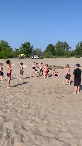 A group of children are playing on a sandy beach on a sunny day. The children are wearing swim trunks and are engaged in what appears to be a game of football. One child is on the ground while others are surrounding him. There are trees and a tent in the background, as well as a white vehicle. The sky is clear and blue.