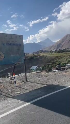 The video shows a scenic mountain landscape under a partly cloudy sky. In the foreground, a person is sitting on the edge of a hill, facing away from the camera. Beyond them, a village with houses and buildings is nestled in the valley, with terraced hillsides and sparse vegetation. Mountains rise in the background, with a prominent peak visible in the distance. A road with a yellow and black striped guardrail runs along the edge of the frame in the lower portion of the video. The sun is shining brightly, casting long shadows. The overall impression is one of a rural, mountainous environment in daylight.