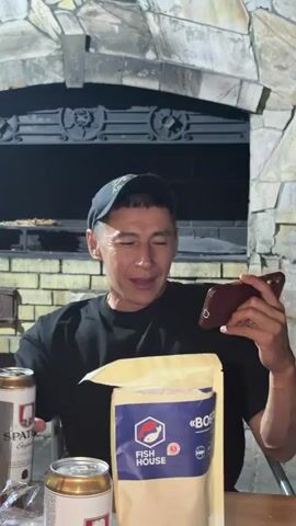 A man wearing a black t-shirt and a baseball cap sits at a table with two cans of beer and a bag of "Fish House" branded product. He is holding a smartphone in his right hand and appears to be looking at the screen with a smile. The background suggests an outdoor setting with a stone structure, possibly a grill or fireplace. The man's left hand is resting on the table.