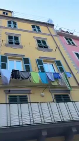 The camera pans up the colorful facades of buildings in a narrow street in a village. The buildings are three and four stories tall, and painted in pink, yellow, and white. They have dark green shutters on the windows. Laundry hangs from a line strung across the front of one of the yellow buildings. Some of the buildings have small balconies. People can be seen walking in the street. A string of decorative lights hangs across the street. The sky is blue.