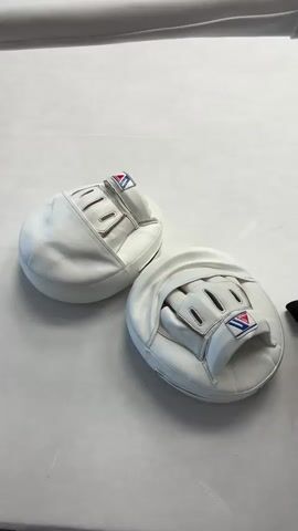 The video shows a pair of white boxing mitts on a white surface. The mitts are oval-shaped with a pocket for the hand, and they have the brand "Winning" on them. The camera then pans up to reveal a boxing ring. The ring is white with black ropes. There are signs on the ropes that say "FITNESS" and "FIGHT TECH". In the background, there is gym equipment, including weight machines and punching bags. The lighting is bright and artificial.