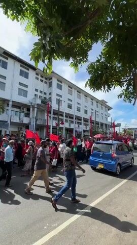 The video shows a large group of people marching down a street. They are carrying red flags and walking on the road, disrupting traffic. The crowd is diverse in age and clothing, with some wearing traditional attire and others in more casual wear. A large white building flanks the street, and trees provide some shade. The sky is blue, indicating a clear day. A blue car is waiting in the road. The scene suggests a political protest or demonstration.