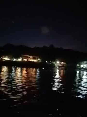The video shows a nighttime view of a body of water, possibly a lake or bay, with lights reflecting off the surface. On the opposite shore, there are buildings or structures with visible lights. The sky is dark with faint cloud cover. The camera appears to be on a moving vessel, as the perspective shifts slightly. An arm and part of a person's shoulder briefly enter the frame on the right side, towards the end of the video.