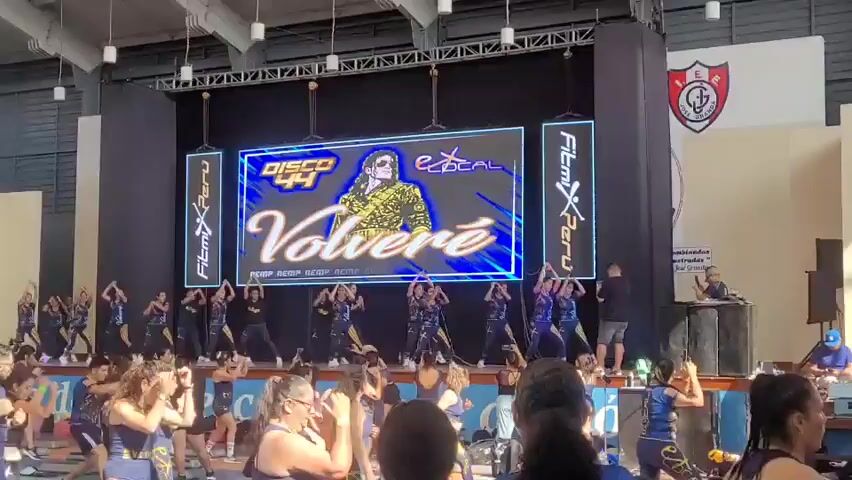 A group of people are participating in an aerobics class or dance event indoors. A large screen displays the words "DISCO 44 Volvere" and an image of a man in a gold jacket. Several performers in matching outfits are dancing on a stage in front of the screen. A large audience is watching the performance, and many are also engaged in some form of physical activity. The environment appears to be a gymnasium or large hall, with athletic equipment visible in the background. There are also audio equipment setups on the side of the stage.