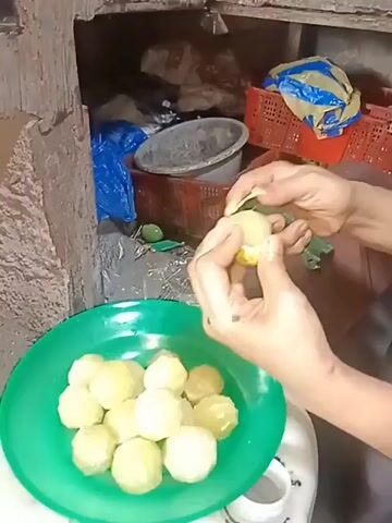 A person's hands are shown preparing food. The hands are peeling small, round, light-colored fruits, possibly potatoes or some type of root vegetable. The peeled fruits are being placed into a bright green plastic bowl. The bowl is filled with many similar peeled fruits. In the background, there are shelves with various items, including red plastic crates, bags, and containers, suggesting a kitchen or market environment. The lighting appears to be artificial and somewhat dim. The person's clothing is partially visible, showing a brown garment. The action is focused on the preparation of the food item.