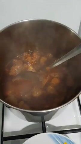 The video shows a large stainless steel pot on a white stovetop, filled with a brown stew. A metal spoon is used to stir the stew, which consists of small pieces of meat in a thick sauce. Steam rises from the pot. There is a glimpse of a blue and white patterned plate near the bottom of the frame. The stovetop and surrounding countertop are white.