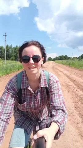 A young woman with dark curly hair, wearing sunglasses and a plaid shirt over a backpack, smiles and talks while riding a bicycle down a dirt road. The road is lined with green fields and trees, and the sky is blue with white clouds. The camera is mounted on the handlebars of the bicycle, providing a first-person perspective of the ride.