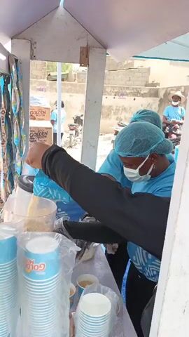 Two women wearing blue shirts and hairnets stand behind a small stand with a white roof. One woman is wearing a black long-sleeved shirt. They are both wearing face masks. The woman in the black shirt holds a blue package and examines it. A third woman in a blue shirt is visible in the background, sitting on a motorcycle. Behind the stand are several boxes labeled "GOLDEN MORN" and hanging packages of the same product. In front of the women are containers of ingredients and stacks of blue and white cups labeled "GOLDEN MORN." The women appear to be preparing food or beverages. The environment is sunny and there is a building in the background.