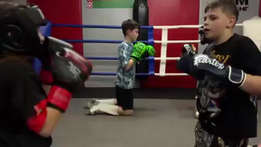 Two young people wearing boxing gear are sparring in a boxing ring. The person facing the camera is wearing black and gold boxing gloves, a black t-shirt, and Muay Thai shorts. The other person is wearing a head guard, black gloves with red hand wraps, and a black t-shirt with writing on the back in a foreign language. Other children are in the background also practicing. The boxing ring has a gray floor, white ropes, and red corners. There is boxing equipment in the background.