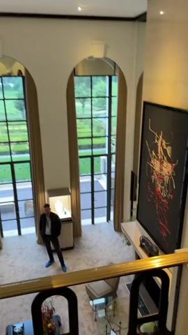 The video showcases the interior of a luxurious, high-ceilinged room, possibly a showroom or a large living space in a mansion. The room features large arched windows that offer a view of an outdoor garden or landscape. A man dressed in dark clothing and blue footwear stands in the center of the room, apparently observing the surroundings. Display cases, which presumably hold jewelry or other valuables, are strategically placed throughout the room. A large circular mirror is mounted on the wall. Additional furniture includes a gray sofa and a chair. The floor is covered with a patterned carpet. A large abstract painting adorns the wall near a fireplace. The camera appears to be positioned on an upper level, providing an overhead view of the room. A person wearing a green hat is seated near the bottom right corner of the frame, possibly filming or taking notes. The general atmosphere is elegant and sophisticated.