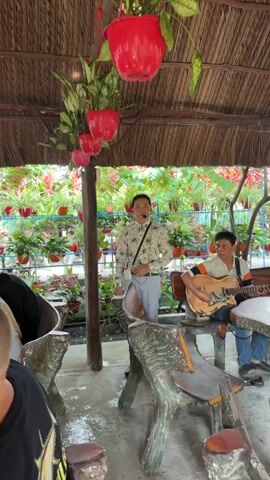 In a vibrant, garden-like setting, two men are engaged in a musical performance. One man, wearing a patterned long-sleeved shirt and light-colored trousers, stands and sings into a headset microphone, with a small white device clipped to his belt. He appears to be the lead vocalist. To his right, another man, dressed in a striped polo shirt, sits on a wooden stool and plays an acoustic guitar.

The scene is semi-outdoor, possibly under a thatched roof structure, indicated by the straw-like material above. The background is filled with an abundance of potted plants, including red flowering varieties and various green foliage, arranged on shelves and hanging from the ceiling in red planters. The overall atmosphere suggests a relaxed and natural environment, likely a cafe, restaurant, or tourist attraction in Thới Sơn, Vietnam, where live traditional or folk music is performed. The lighting indicates it is daytime.