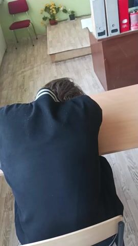 The video shows a school classroom. In the initial frame, a person is resting their head on a desk. The camera then pans to show backpacks on the floor and on a desk. Several students are seen sitting at desks, each looking at their cell phones. The classroom has wooden desks, chairs, and cabinets. Potted plants are placed on the cabinets. The floor is covered with light-colored wood-like planks.