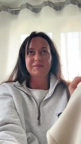 A woman with long dark hair and bangs, wearing a grey zip-up sweatshirt over a white t-shirt, looks upwards and to her right. Her right hand is raised, holding a clear wine glass filled with a pale yellow liquid. She appears to be smiling slightly. The background consists of a window with white curtains and grey pelmets. The camera is positioned below her, looking up at her face.