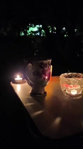 A close-up, low-angle shot shows a table with a cup and two tea light candles in holders. The cup is white with purple flowers and a gold trim. The tea light on the left is in a small white holder. The tea light on the right is in a clear, hexagonal glass holder. The cup is steaming, suggesting it contains a hot beverage. The table appears to be made of wood and is a light color. The background is dark and out of focus, with trees and some blurry lights visible. The scene is illuminated by the light of the candles.