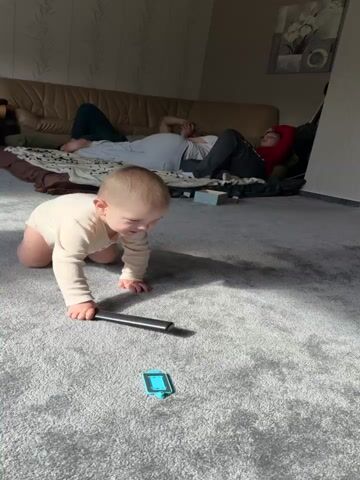 A video shows a baby crawling on a gray carpet in a living room. The baby is wearing a light-colored long-sleeved onesie. The baby crawls toward a black cylindrical object and a small blue object, moving its hands and knees in a coordinated motion. In the background, two adults are visible on a beige sofa, seemingly resting. One adult is lying down, and the other is sitting up. The room has light-colored walls, and a wooden door is visible to the right. Various household items and baby gear, such as a high chair, are also in the background.