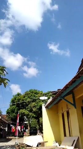 The video opens with a view of a bright blue sky with scattered white clouds. In the foreground, a yellow building with a tiled roof is visible on the right. To the left of the building, trees and some houses can be seen. A flag is also present, hanging vertically. The scene appears to be outdoors in a rural or semi-rural setting. There are no people or animals clearly visible in the frame. The overall lighting suggests it is daytime, likely under sunny conditions.