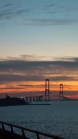 The video shows a wide shot of a bridge spanning a body of water under a colorful sunset sky. The sky transitions from a pale blue at the top to orange and red near the horizon, with dark, streaky clouds in between. The bridge, a suspension bridge with two prominent towers, is silhouetted against the bright sky, with its lights visible along the span. In the foreground, a dark railing is visible, suggesting the shot is taken from a moving vessel or platform. A small landmass with a lighthouse is situated to the left of the bridge. A lone bird flies across the sky in the initial seconds of the video. The water is dark and slightly rippled. The camera remains static throughout the video.