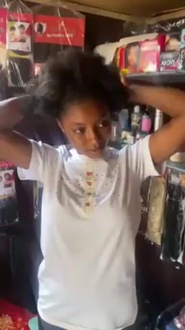 A woman with dark hair and skin, wearing a white t-shirt with a yellow stripe down the middle, stands in what appears to be a small shop. She is holding her hair up with both hands. Shelves behind her are stocked with hair products and wigs. The shop has a cluttered, informal look.