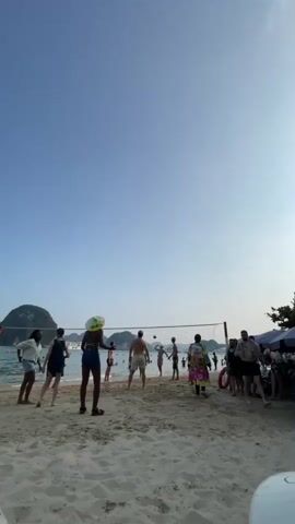 A group of people are playing volleyball on a sandy beach. The sky is clear and blue. In the background, there are islands and boats. The people are of various ages and ethnicities. They are dressed in casual beach attire. The volleyball game appears to be friendly.