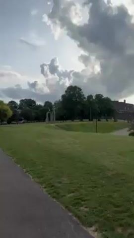 The camera pans across a grassy park on an overcast day. In the distance, there is a large, brick building surrounded by trees. Closer to the camera, there is a series of archways made of light-colored stone. The grass is green and well-maintained. There is a road visible in the middle of the frame. The sky is cloudy.