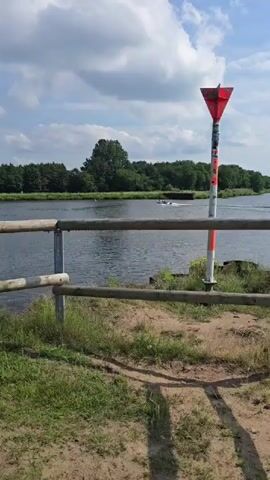 The video opens with a view of a body of water, possibly a river or canal, with a wooden fence in the foreground. A red and white navigation marker is visible on the right side of the frame. Trees line the opposite bank of the water. In the distance, a small boat or raft can be seen on the water. The sky is partly cloudy with a bright blue sky. The camera pans slowly across the scene, showing the water, the fence, and the distant shore. There are no people visible in the video. The video appears to have been filmed on a sunny day.