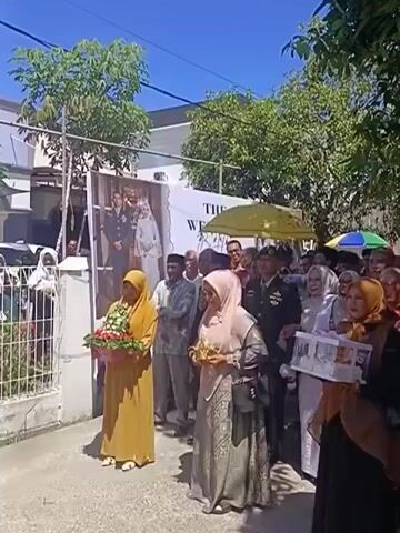 A group of people, predominantly women wearing headscarves and traditional Islamic attire, are gathered outdoors on a sunny day. Some are holding flowers and small boxes, possibly gifts or offerings. In the background, a large banner displays a wedding photograph of a couple in formal attire. A few men, some in military uniform, are also present among the crowd. The scene appears to be a procession or a welcoming ceremony, with people lining a pathway. Trees and buildings are visible in the background, suggesting an urban or suburban setting.
