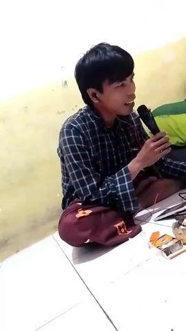 A man sits cross-legged on the floor, holding a microphone to his mouth and wearing earbuds. He is dressed in a plaid shirt and maroon pants. To his right, on the floor, are a cigarette pack and a lighter. The background is a plain wall.