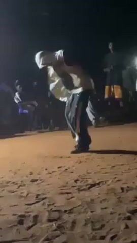 The video shows a man dancing in a dirt field at night. He is wearing a white shirt, dark pants, and has a white cloth covering his head. Several people are watching him dance in the background, some sitting on chairs. The scene is dimly lit, and the background is blurry, but it looks like there are some houses or buildings nearby. The camera is handheld and slightly shaky, and it moves around to follow the dancer.