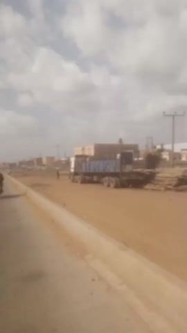 The video shows a road with a truck parked on the side. The sky is partly cloudy. A person walks across a dirt area next to the road. A motorcycle passes on the road. The video pans along the road. Buildings and power poles are visible in the background.