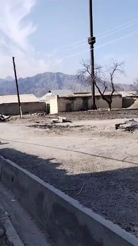 The video presents a view of a rural or semi-rural street on a sunny day. The road is unpaved and appears to be a mixture of dirt and gravel. Along the roadside is a concrete curb.  Several low buildings, possibly houses or sheds, line the street, with mountains visible in the distance. There are bare trees scattered throughout the area, as well as utility poles with power lines. Debris or construction materials are lying on the ground in some areas.