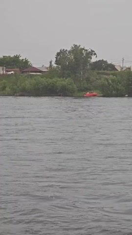 A wide shot of a body of water with a cloudy sky. The shore is lined with trees and bushes. On the shore, there is a red boat and a white object that could be a car. There are also houses and power lines visible in the distance. The water is choppy and gray. The overall impression is one of a gray, overcast day.