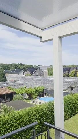 The video pans across a backyard from a balcony. In the foreground, a metal railing with glass panels is visible. The backyard features a green lawn, a hedge, a small gazebo with a table and chairs, a small statue, and a tree. Beyond the yard, there are several greenhouses or similar structures with tiled roofs, followed by houses and more trees under a clear blue sky. A swimming pool is partially visible in the left side of the frame.