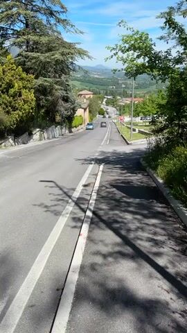 The video presents a series of daytime shots capturing scenic roads and landscapes in Camerino, Italy.

The first two shots (0:00-0:02) offer a wide view down a paved, downhill road. To the left, a large evergreen tree stands prominently, with a building featuring a red-tiled roof visible below it. The road, marked with white lines, descends into a valley, revealing distant green hills and mountains under a clear blue sky with sparse clouds. Several cars are visible on the road in the distance.

The third shot (0:02-0:03) focuses on a more immediate roadside view, showing dense green foliage and trees casting shadows on a paved surface. A vehicle is partially obscured by the greenery on the left, and a signpost or structure is embedded within the plants on the right.

The final shot (0:03-0:04) shows a paved road curving to the right. A black car is parked on the left side of the road, next to a hedge and a white informational sign (text not clearly legible). In the background, houses with red roofs are visible, along with construction cranes against the bright blue sky.

Throughout the video, the weather is sunny and clear, casting distinct shadows and highlighting the vibrant green of the foliage. The overall impression is that of a pleasant drive or walk through a hilly, semi-rural area with elements of urban development. Spoken Italian can be heard in the background.