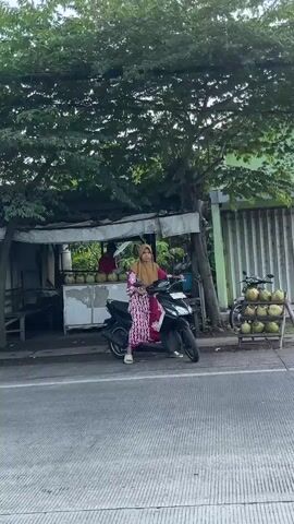A woman wearing a pink and white patterned dress and a hijab is sitting on a black scooter. She is positioned on the right side of the street, in front of a small roadside stall. The stall has a white counter with coconuts on display and a tarp covering the top. Behind the stall, a person in a red shirt is partially visible. To the right of the stall, a wooden rack holds more coconuts. A bicycle is parked next to the rack. The scene appears to be outdoors with trees providing shade over the stall and the street.