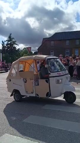 A white, three-wheeled vehicle with a tan canopy is driving down a street. The vehicle has two doors on the side and a covered passenger area. A man is driving the vehicle. Several people are standing on the sidewalks, watching the vehicle go by. Some people are holding phones up, possibly taking pictures or videos. The sky is cloudy. The environment appears to be a town or city street with buildings visible in the background. The vehicle has some writing on the side, including "Best In Klaipėda". The vehicle also has a license plate.