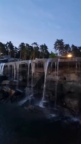 The media file captures a serene natural scene at dusk. The primary subject is a man-made waterfall cascading over rough, dark rocks. The water forms thin, elongated streams as it falls, creating a gentle visual effect. Above the waterfall, a line of tall pine trees with dark green foliage stands silhouetted against a pale blue sky that transitions to a deeper blue towards the top, indicating twilight. The ground surrounding the waterfall appears rocky and uneven, with some larger boulders visible.

A subtle light source, possibly a lamp or building light, is visible in the upper right portion of the frame, casting a warm glow. The overall atmosphere is peaceful and tranquil, amplified by the fading light and the natural sound of the falling water. The time of day is clearly evening, as evidenced by the twilight sky and the soft lighting. The location cues, such as the pine trees and the rugged terrain, combined with the provided location context of Razdory, Russia, suggest a park or recreational area within that region. No people are visible in the frame, and there is no discernible text. The activity is the continuous flow of the waterfall, a natural process enhanced by human landscaping.
