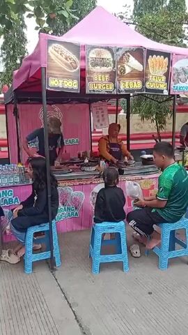 The video shows a street food stall with a pink canopy. The stall has a sign showing the foods it sells: hotdog, beef burger, kebab, "telur gulung" (egg roll), and takoyaki. Two people are working at the stall preparing food. Three customers are sitting on blue plastic stools in front of the stall eating. A man in a green shirt is eating from a container, a girl in a black jacket is also eating, and a woman in a black shirt is sitting on the stool. There are bottles of water on the counter. The scene is outdoors with trees in the background.
