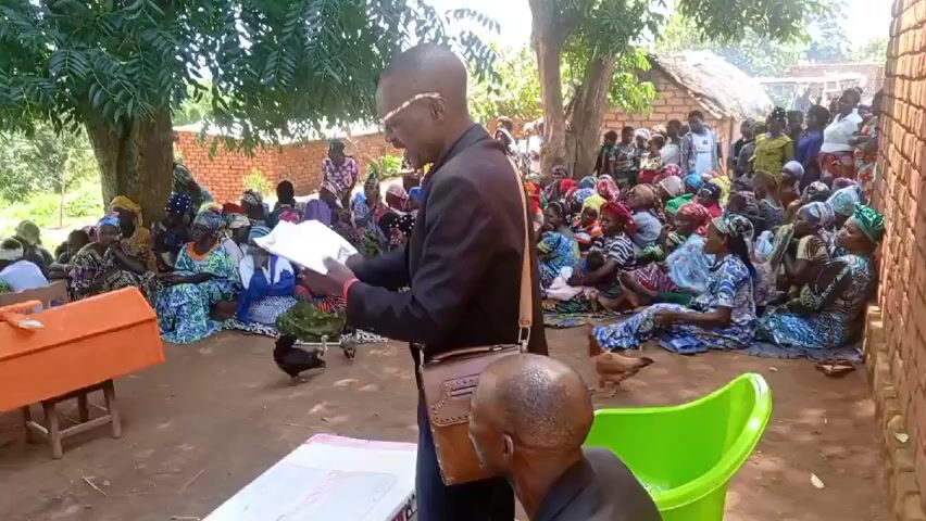 Here's a description of the video:

The video shows a funeral taking place outdoors in what appears to be a rural African setting. A large group of people, mostly women, are gathered on the ground, many wearing colorful headscarves. A man in a suit and glasses stands in front of the group, reading from a book. An orange coffin rests on a simple wooden frame. A few chickens wander around the gathering. A man sits on a green chair nearby, listening. 

A brick building and trees are visible in the background. There is singing and chanting. The atmosphere seems solemn. One man is kneeling behind the coffin. Another man appears to be lying on the ground near the coffin.