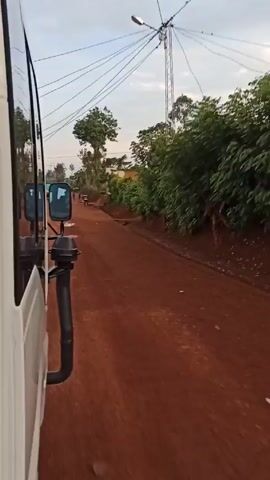 The video shows a view from the side of a white vehicle driving on a red dirt road. Power lines and a communication tower are visible in the sky. Along the road, there are bushes, trees, and a few simple buildings. Several people are walking in the distance along the road. The sky is light blue with some clouds. The road surface is uneven with puddles of water. The camera is mostly static, capturing the motion of the vehicle.