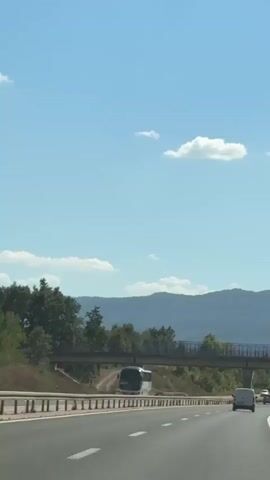 The video shows a highway scene on a clear day. The camera is positioned inside a moving vehicle, looking forward. The road is paved with asphalt and has white lane markings. Several vehicles are visible on the highway, including a bus, vans, and trucks. A pedestrian overpass spans the highway in the middle ground. In the background, there are rolling hills covered with trees, and the sky is blue with a few scattered white clouds. The perspective suggests the viewer is traveling at highway speed.