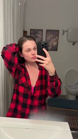 A woman stands in a bathroom and takes a selfie with a black cell phone. She wears a red and black flannel shirt over a pink top. She is touching her brown hair with her right hand. Behind her is a white sink and mirror. To the right are two framed pictures, a white toilet with the lid up, and a blue cushioned bench. A white shower curtain hangs to the left of the mirror.